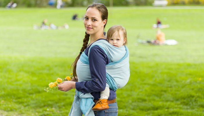 59239832 - a young mother carrying her little baby in a sling on her back and walked in beautiful sunny weather by a park. the two laugh and have fun